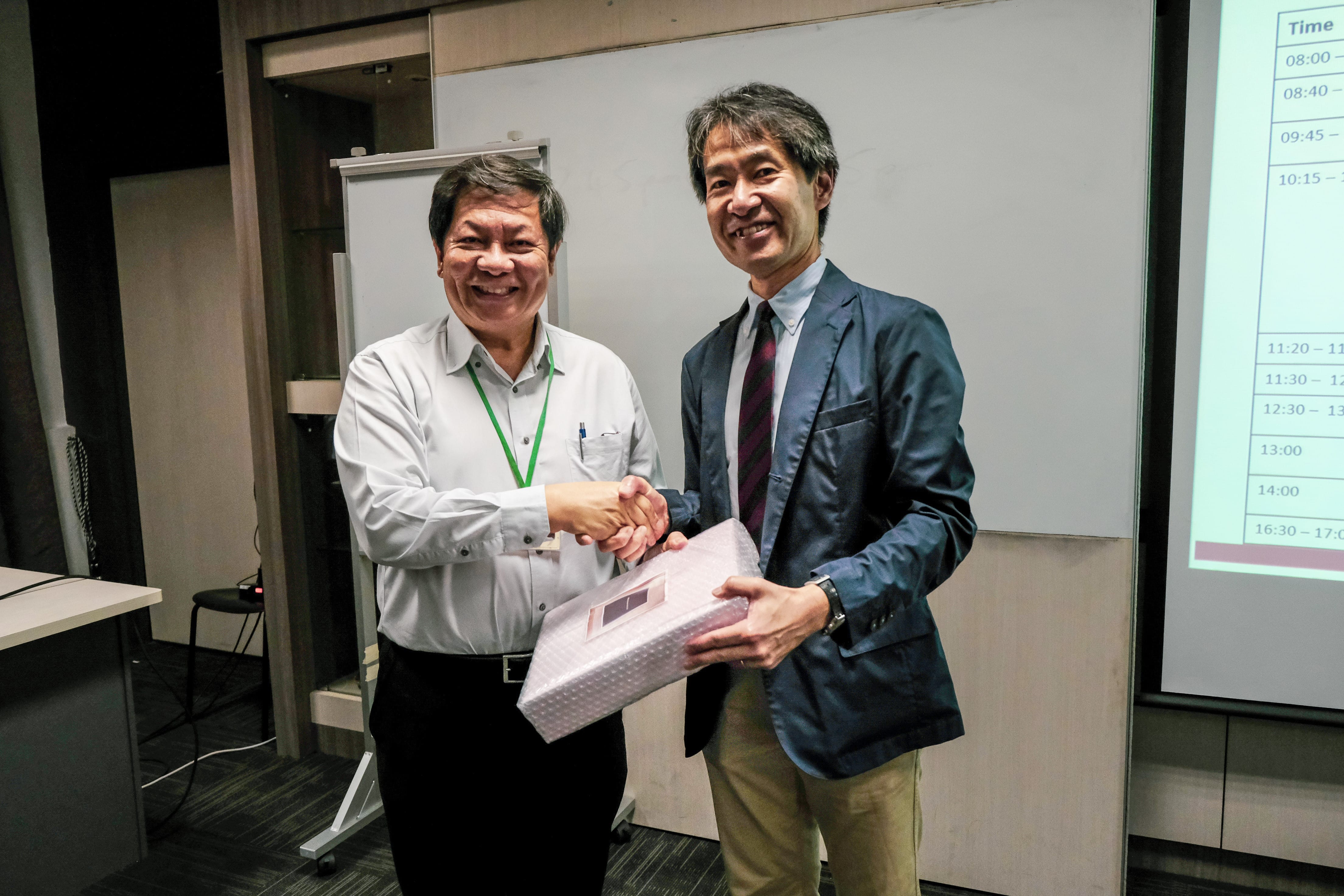 Two smiling men shake hands while holding a wrapped gift in a conference room with a projected schedule.